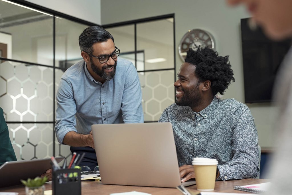 Portrait of two casual business partners sitting at table together and working. Group of creative businesspeople working together at modern office. Handsome mature businessman discussing affairs with African man and smiling together in a meeting room.